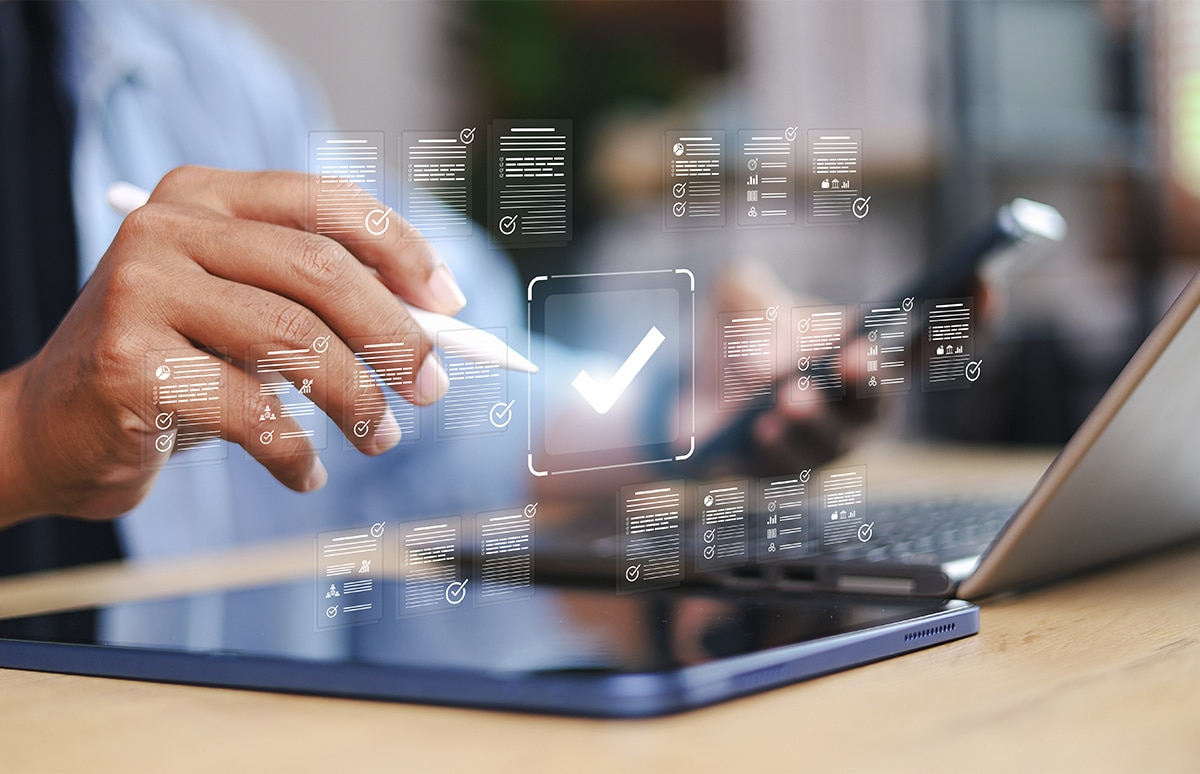 Mano interactuando con una tableta digital mediante un lápiz táctil, con una interfaz virtual superpuesta que muestra listas de verificación y un icono central de validación, junto a un ordenador portátil sobre una mesa de trabajo