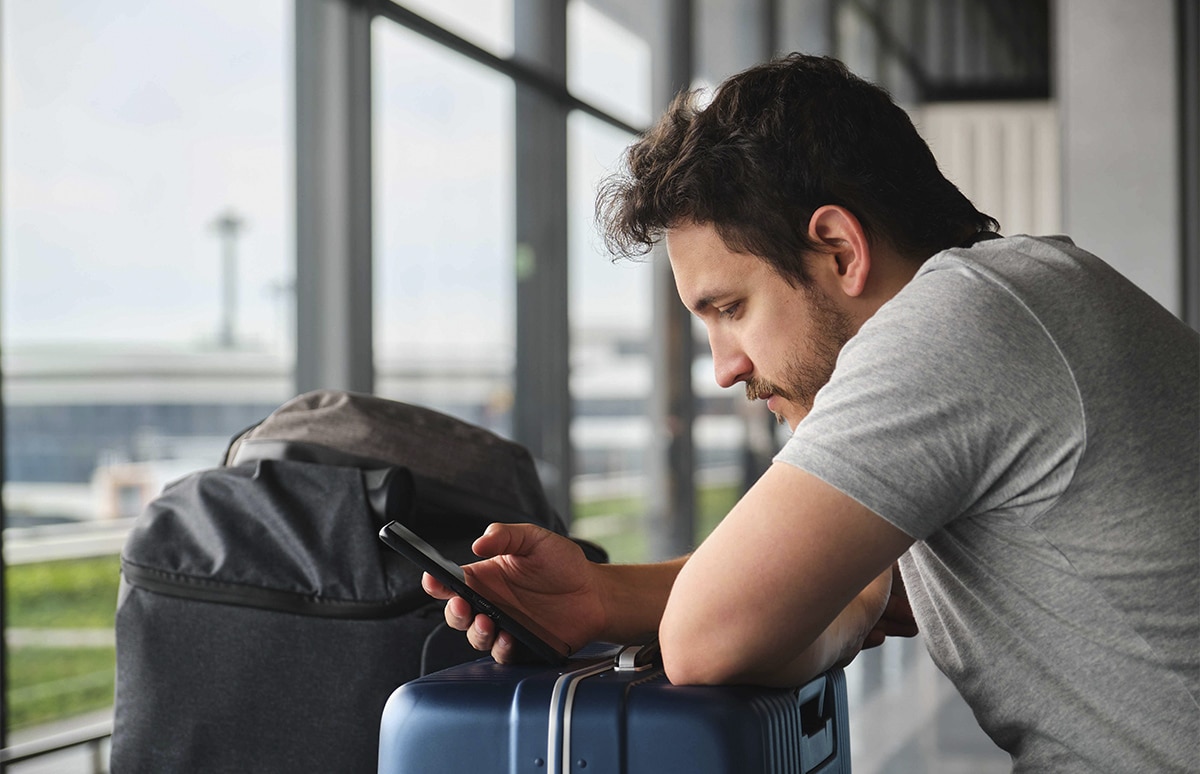 Persona apoyada sobre equipaje en una terminal de transporte, consultando un teléfono móvil junto a grandes ventanales