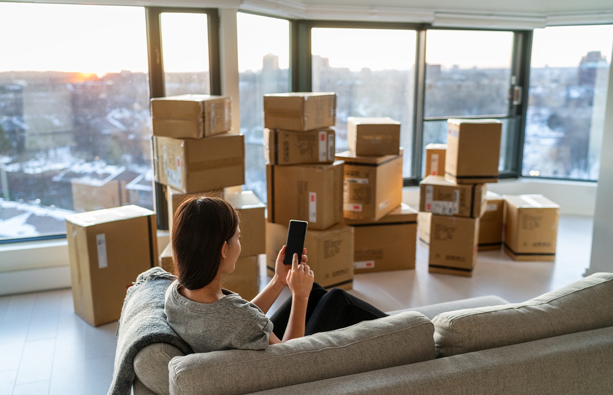 Mujer mirando un móvil sentada en un sillón en una habitación vacía con cajas de mudanza