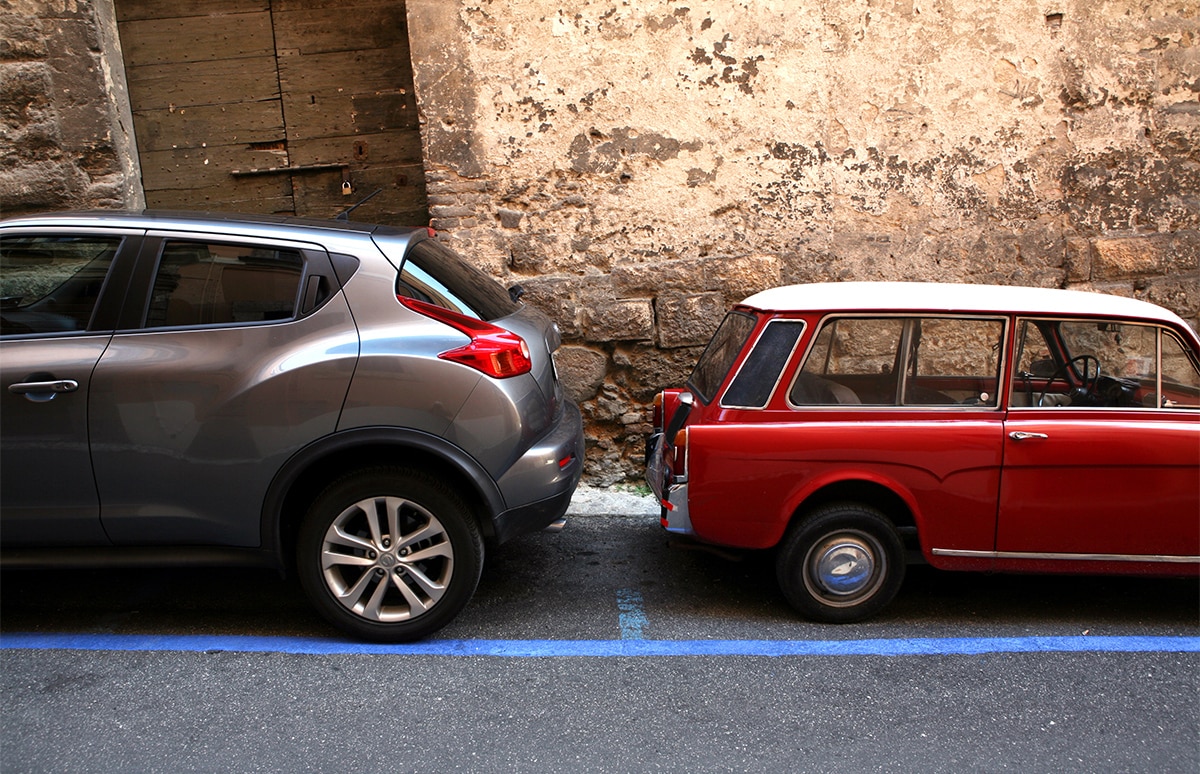 Dos coches estacionados muy próximos entre sí —uno moderno gris y otro clásico rojo— junto a una pared de piedra envejecida