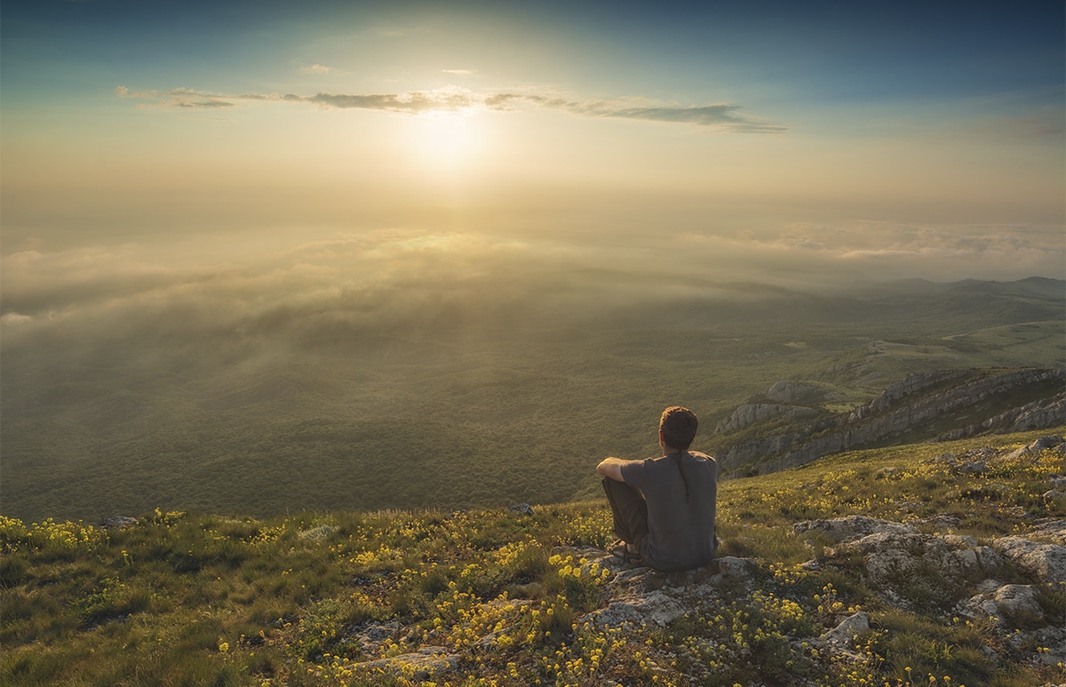 Persona sentada en lo alto de una montaña contemplando un paisaje iluminado por el sol