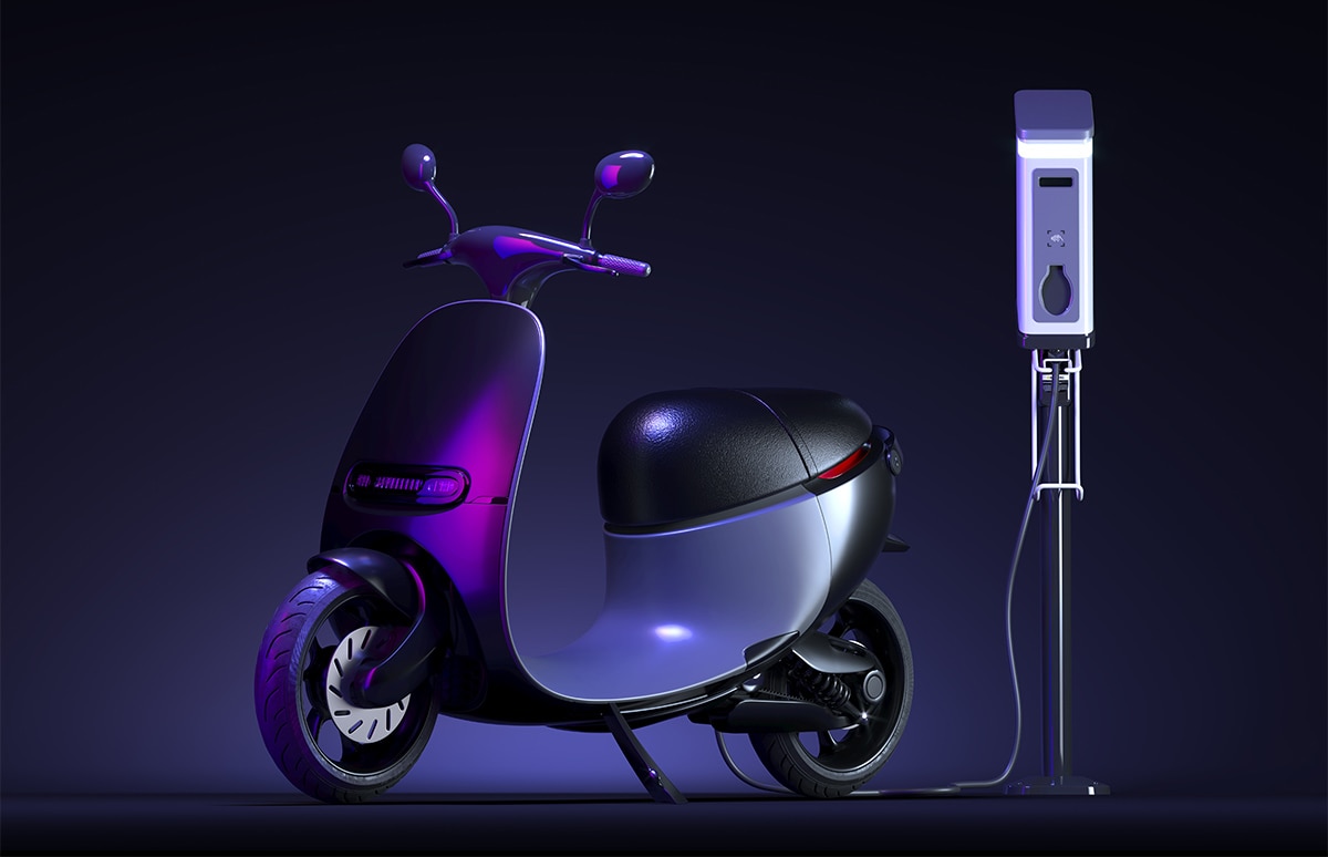 Motocicleta eléctrica conectada a un punto de recarga mediante cable, presentada en un entorno oscuro con iluminación artificial que resalta el diseño y el sistema de carga