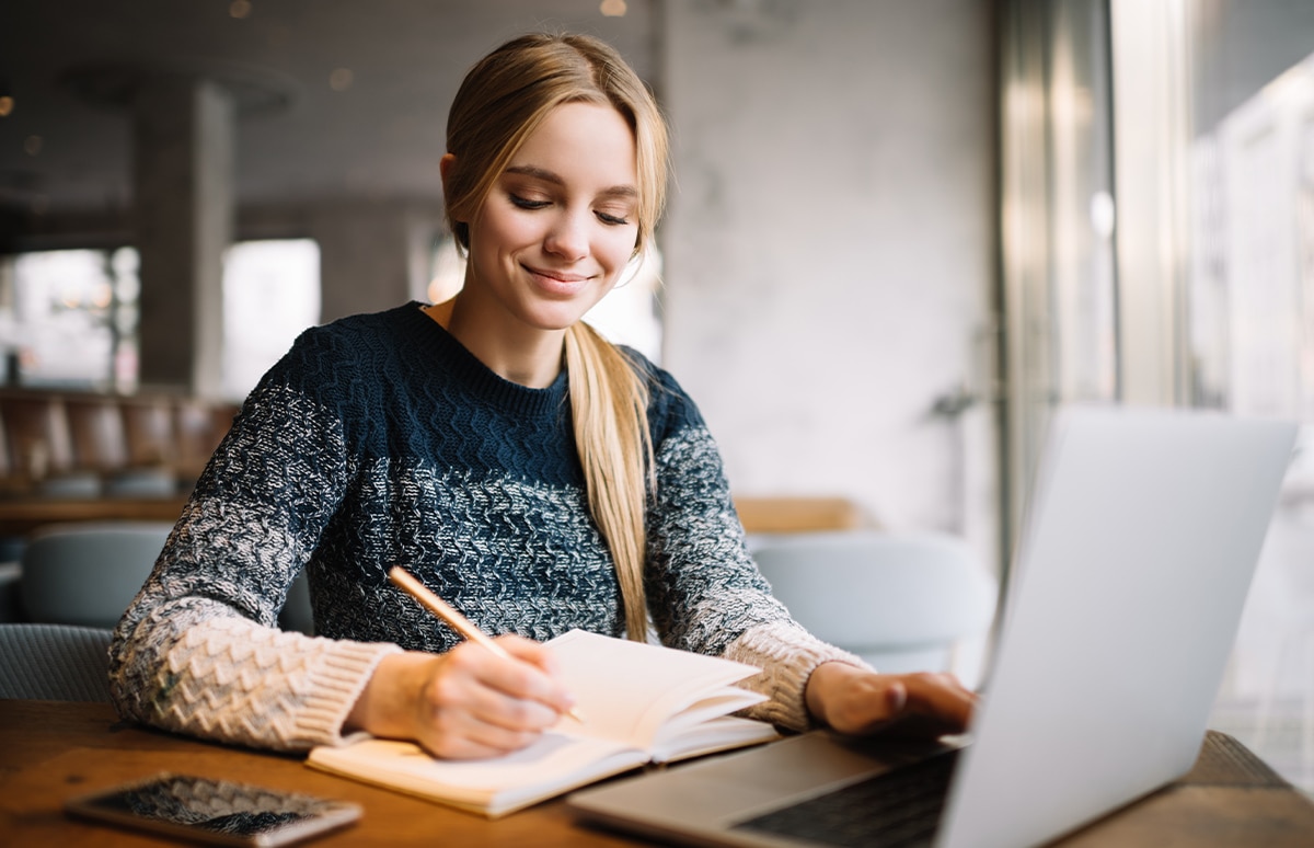 Mujer estudiante sonriente escribiendo en un cuaderno, con un portátil enfrente