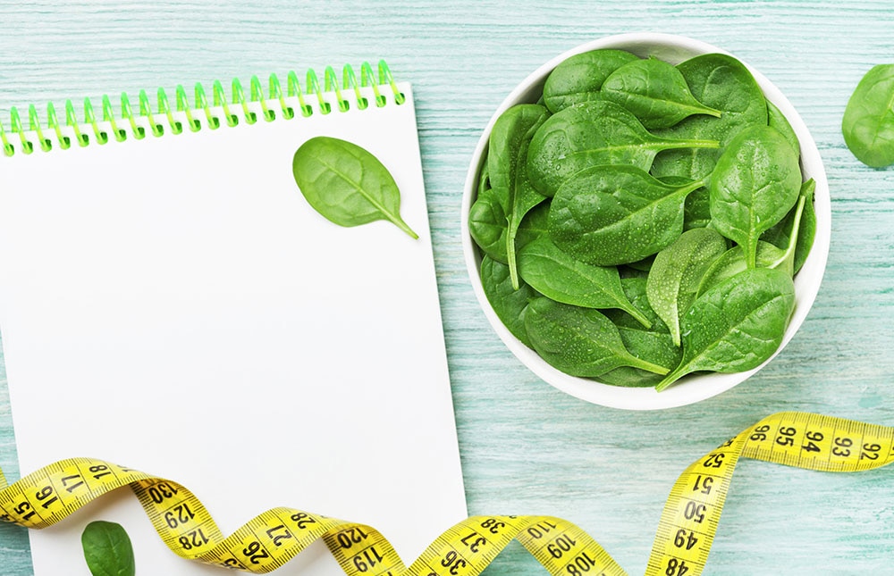 Notebook, green spinach leaves and tape measure on wooden table top view. Diet and healthy food concept.