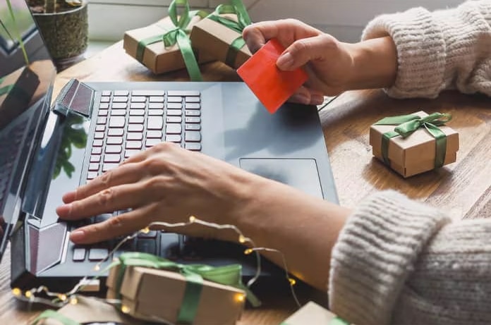 Manos realizando una compra online con una tarjeta sobre un portátil rodeado de pequeños paquetes de regalo