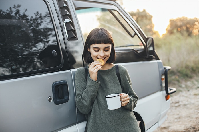 Chica con una taza en su mano comiendo una galleta al lado de una furgoneta