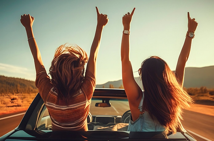 Dos personas disfrutan de un viaje en coche descapotable al atardecer, levantando los brazos con alegría mientras avanzan por una carretera rodeada de paisaje natural
