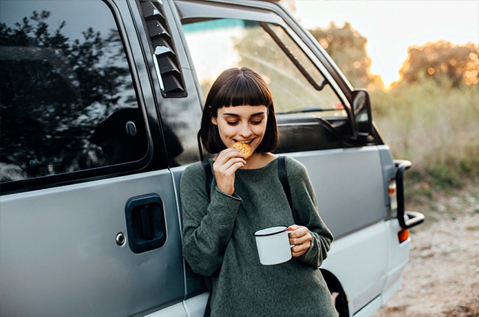 Mujer joven apoyada en una furgoneta, disfrutando de una bebida caliente en un entorno natural