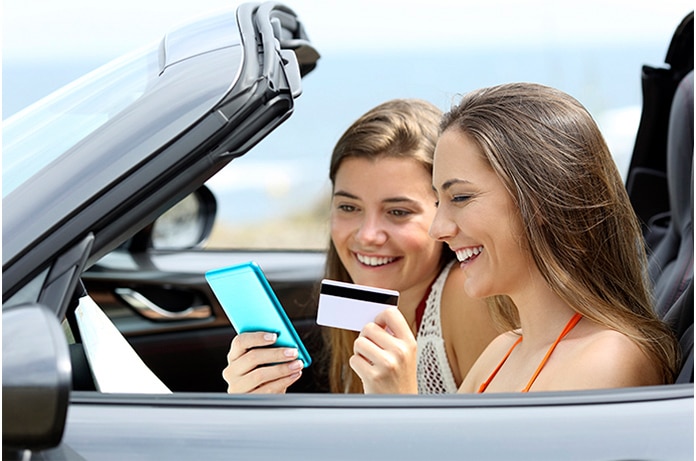 Dos mujeres jóvenes en un coche descapotable realizando una compra online con el móvil y una tarjeta