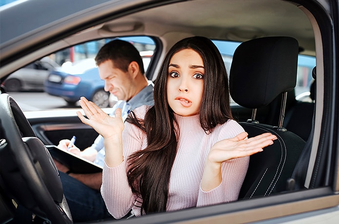Mujer joven al volante gesticulando con las manos mientras un instructor toma notas durante una práctica o evaluación de conducción.