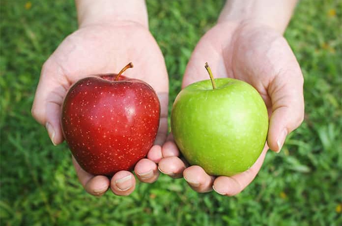 Dos manzanas, una roja y una verde, sostenidas en ambas manos sobre césped, simbolizando comparación entre opciones