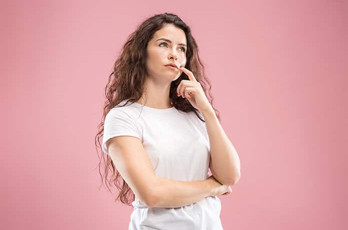 Persona en postura pensativa, con la mano en el mentón y los brazos cruzados, sobre un fondo rosa suave