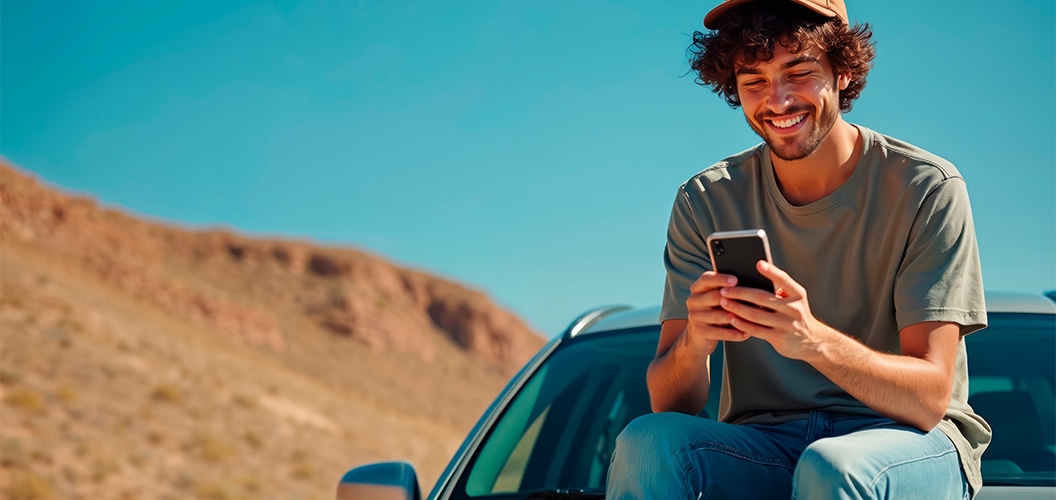 Persona sentada sobre el capó de un vehículo estacionado en un entorno árido, consultando un teléfono móvil bajo un cielo despejado