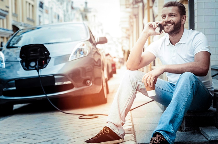 Hombre sentado en una acera hablando por teléfono mientras su coche eléctrico se encuentra conectado a un punto de carga cercano
