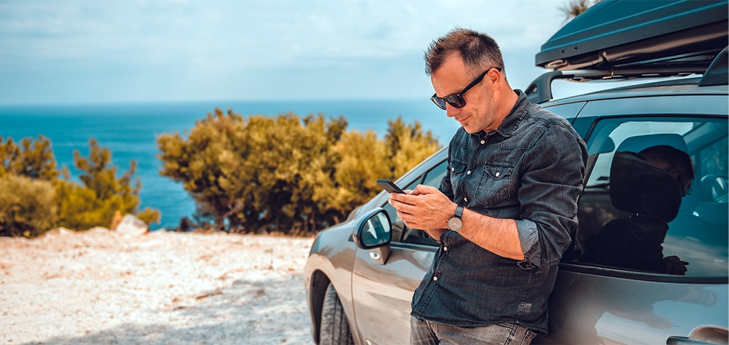 Persona de pie junto a un vehículo con portaequipajes, consultando un teléfono móvil frente a un paisaje costero con vegetación y mar al fondo