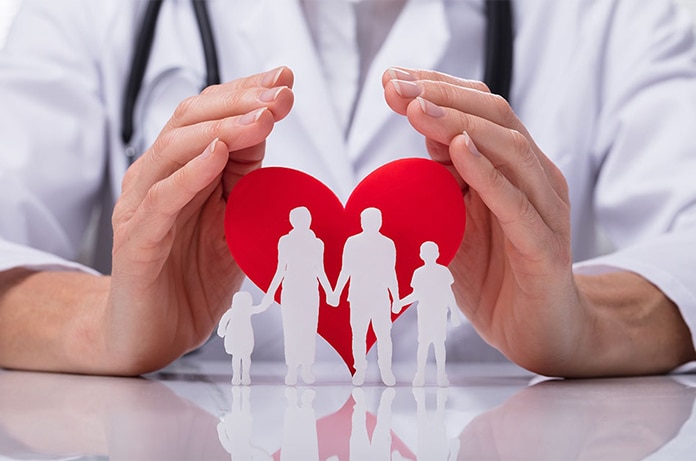 Manos de una persona con bata médica protegiendo la silueta de una familia sobre un corazón rojo, representando cuidado y protección sanitaria