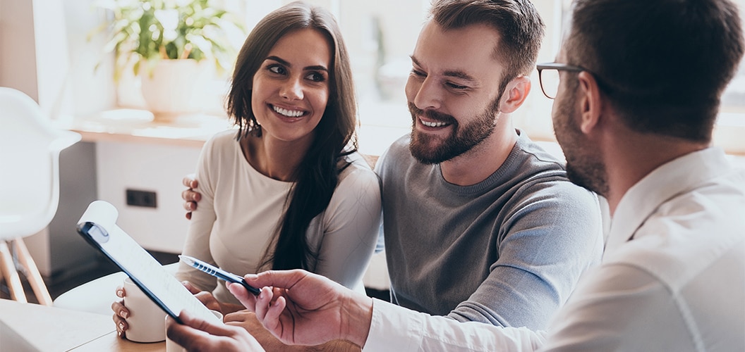 Dos personas sentadas juntas mientras reciben explicaciones de una tercera persona que muestra una carpeta durante una consulta en un entorno interior luminoso