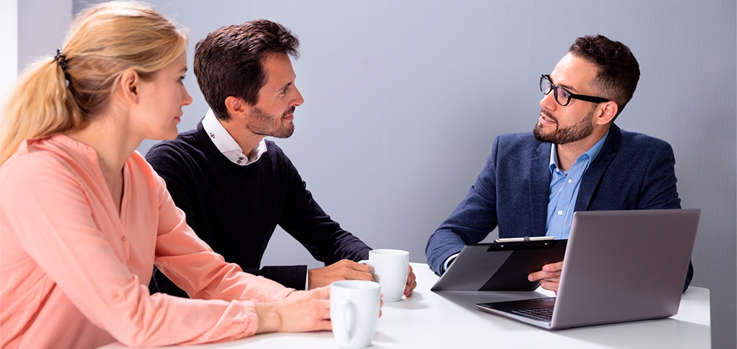 Varias personas dialogando alrededor de una mesa con tazas y un ordenador portátil mientras un profesional toma notas en una carpeta