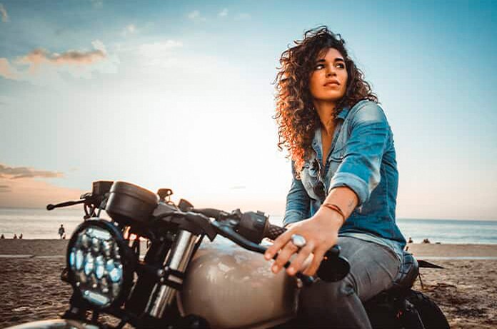 Figura apoyada sobre una motocicleta en la playa al atardecer, con el mar y el cielo iluminado al fondo