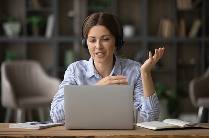 Figura utilizando auriculares con micrófono mientras participa en una reunión virtual desde un escritorio con libros y un ordenador portátil
