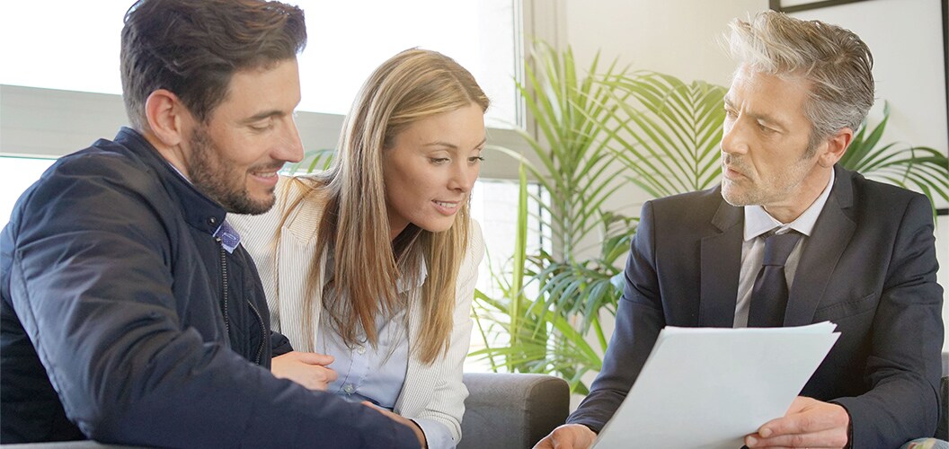 Dos personas consultando documentación que un profesional les muestra durante una reunión en un entorno interior con plantas y luz natural