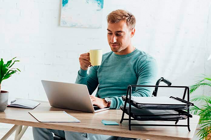 Figura sentada frente a un ordenador portátil sujetando una taza en una estancia iluminada y decorada con plantas