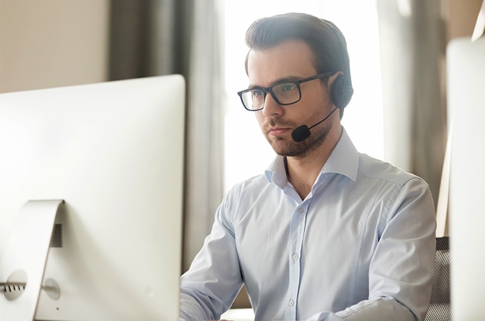 Persona trabajando frente a un ordenador de sobremesa en un espacio interior iluminado, utilizando auriculares mientras consulta la pantalla en un entorno de trabajo ordenado