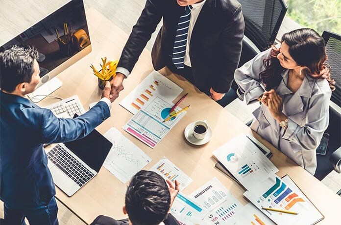 Varias personas reunidas en una mesa con documentos y gráficos financieros, mientras dos de ellas sellan un acuerdo con un apretón de manos