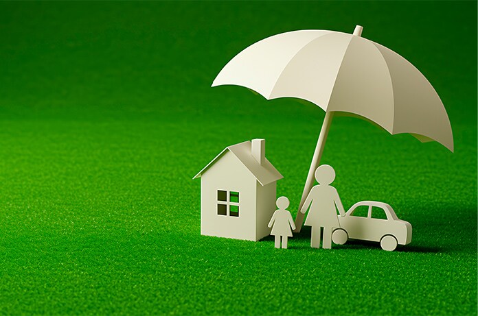 Figura de una casa, una familia y un coche protegidos bajo un gran paraguas sobre una superficie verde