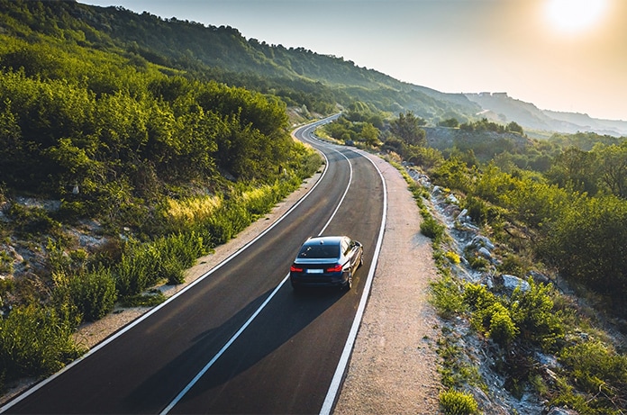 Automóvil circulando por una carretera sinuosa rodeada de vegetación frondosa al atardecer, con el sol bajo en el horizonte