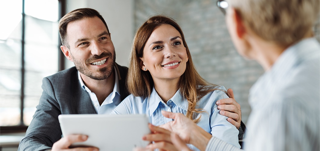 Dos personas sentadas frente a una tercera persona que les muestra información en una tableta durante una reunión en un entorno interior iluminado