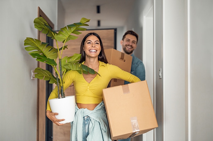Dos figuras transportando objetos durante una mudanza, una de ellas llevando una maceta grande y una caja de cartón por un pasillo interior