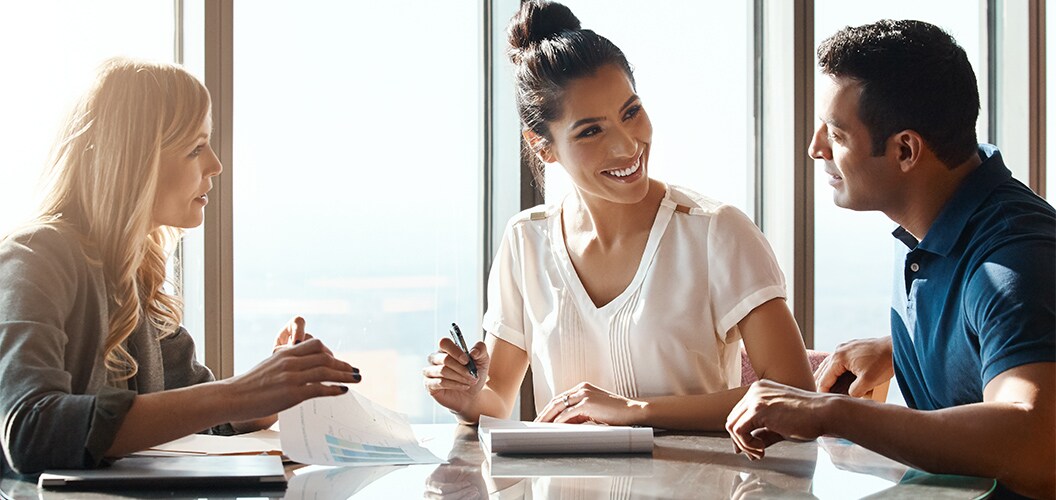 res personas reunidas en una mesa de oficina, revisando documentos mientras una de ellas toma notas con un bolígrafo en un ambiente iluminado por luz natural