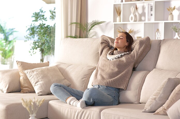 Figura recostada en un sofá amplio con los brazos detrás de la cabeza en un salón decorado con tonos claros y textiles suaves