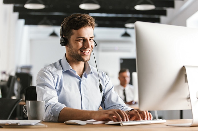 Persona utilizando auriculares y teclado frente a un ordenador de sobremesa en una oficina diáfana con otros puestos de trabajo al fondo