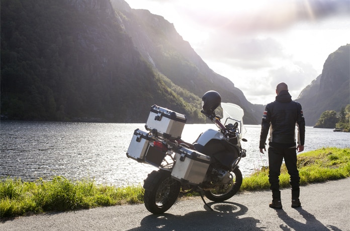 Motocicleta estacionada junto a una carretera junto a un lago, con una persona de pie observando un paisaje montañoso