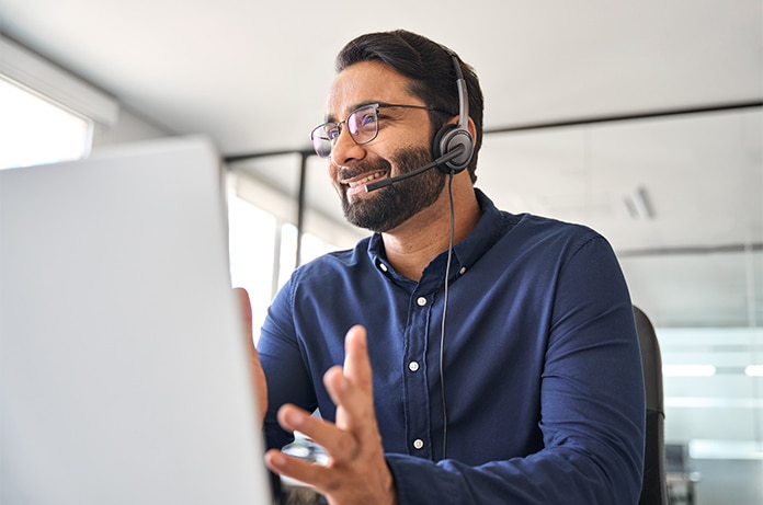 Persona participando en una videollamada desde una oficina acristalada, utilizando auriculares y gesticulando frente a un ordenador portátil