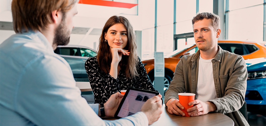 Reunión en un concesionario, con varias personas sentadas alrededor de una mesa mientras una de ellas muestra información en una tableta, con vehículos expuestos al fondo