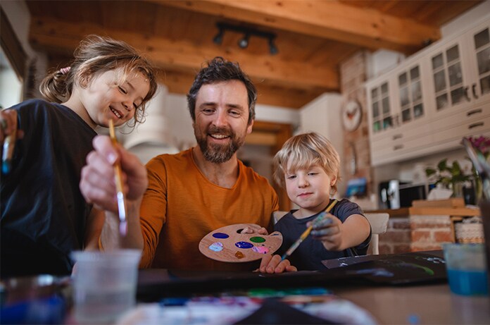 Actividad creativa en un hogar, con personas adultas y una criatura pintando juntos sobre una mesa con pinceles, pinturas y una paleta de colores