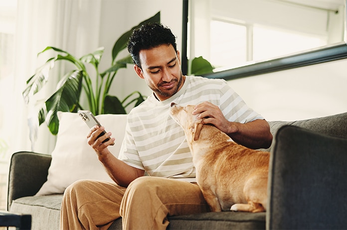 Persona sentada en un sofá utilizando un teléfono móvil mientras acaricia a un animal de compañía en un salón con plantas y luz natural