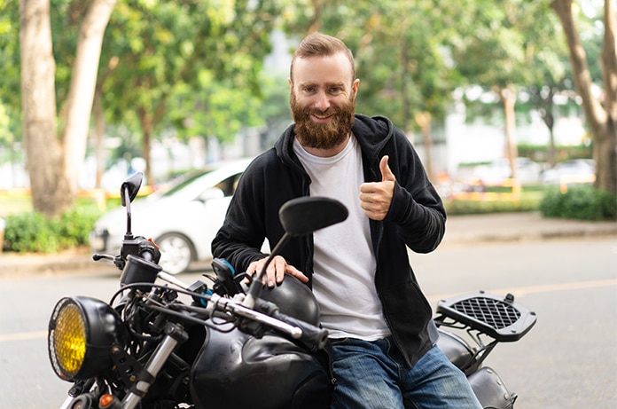 Persona sentada sobre una motocicleta estacionada en una zona urbana arbolada, realizando un gesto de aprobación con la mano