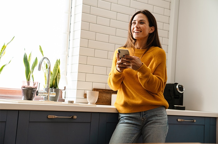 Persona apoyada en una encimera de cocina moderna, utilizando un teléfono móvil junto a plantas y pequeños electrodomésticos