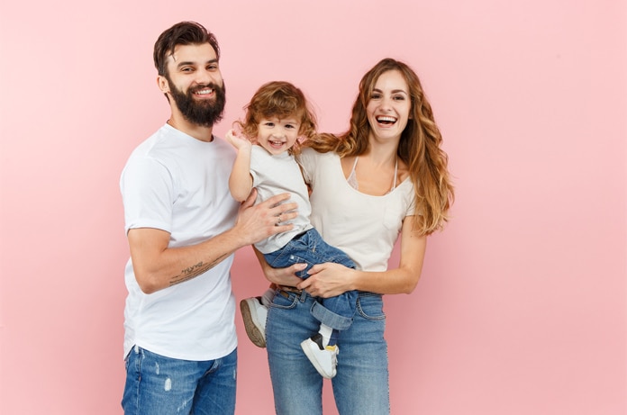 Tres personas posando frente a un fondo liso de color rosa, con una persona adulta sosteniendo a una criatura en brazos, todas vestidas con ropa casual en tonos claros y vaqueros