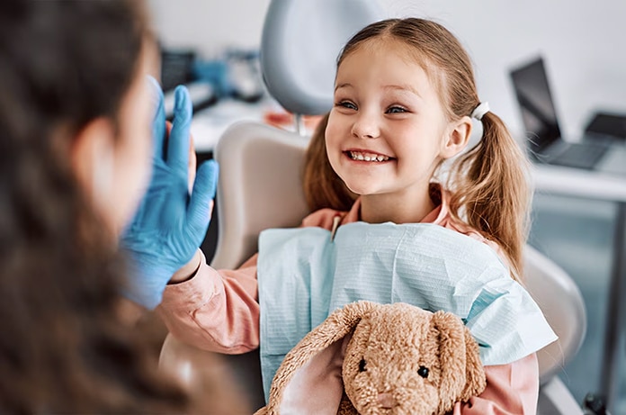 Menor sentada en un sillón dental dando un gesto de felicitación con una mano enguantada enfrente, mientras sostiene un peluche