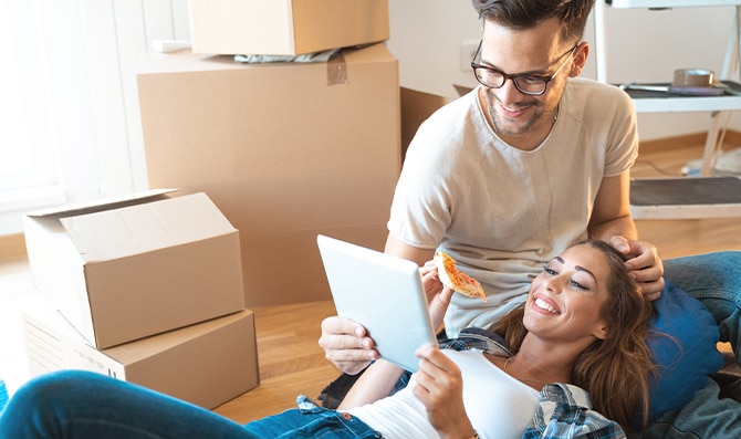 Dos personas entre cajas de mudanza descansando en el suelo mientras revisan una tableta y comparten una porción de pizza