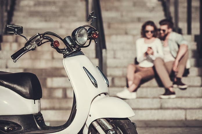 Un ciclomotor blanco estacionado en primer plano con dos personas sentadas en unas escaleras al fondo consultando un teléfono