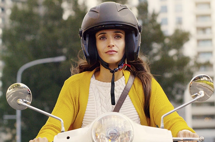 Persona manejando un ciclomotor blanco vestida con una chaqueta amarilla en una zona urbana arbolada