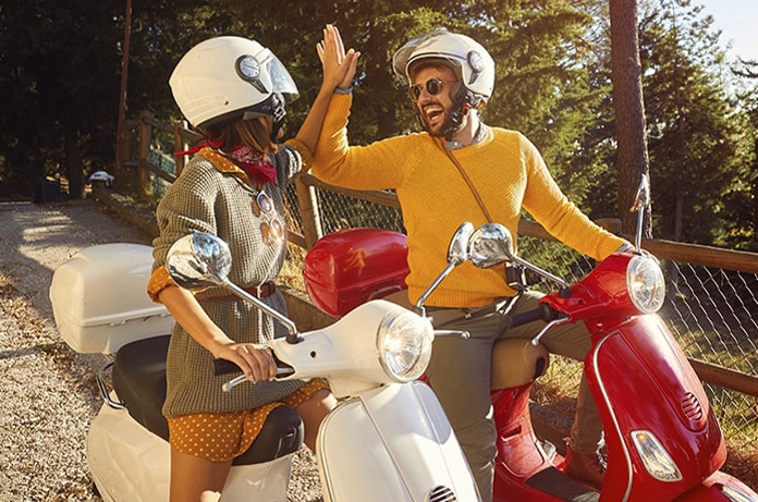 Dos personas con cascos saludándose con un gesto mientras están detenidas sobre sus scooters en un camino arbolado