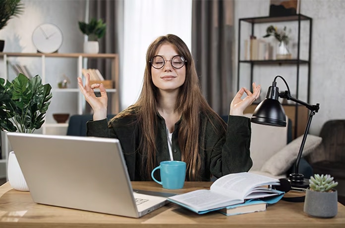 Persona sentada frente a un ordenador portátil realizando un gesto de relajación con ambas manos mientras estudia en un escritorio con libros y una taza