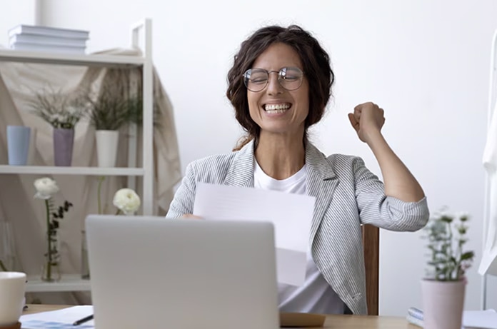 Persona levantando un brazo en señal de logro mientras sostiene varios documentos frente a un ordenador portátil en un espacio de trabajo iluminado